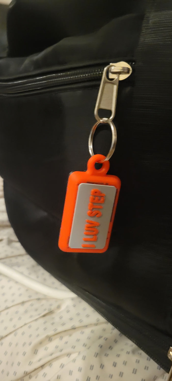 An orange rectangular luggage tag with a silver reflective panel that reads 'I LOVE STEP' in red, attached to a silver key ring and zipper pull of a black bag.
