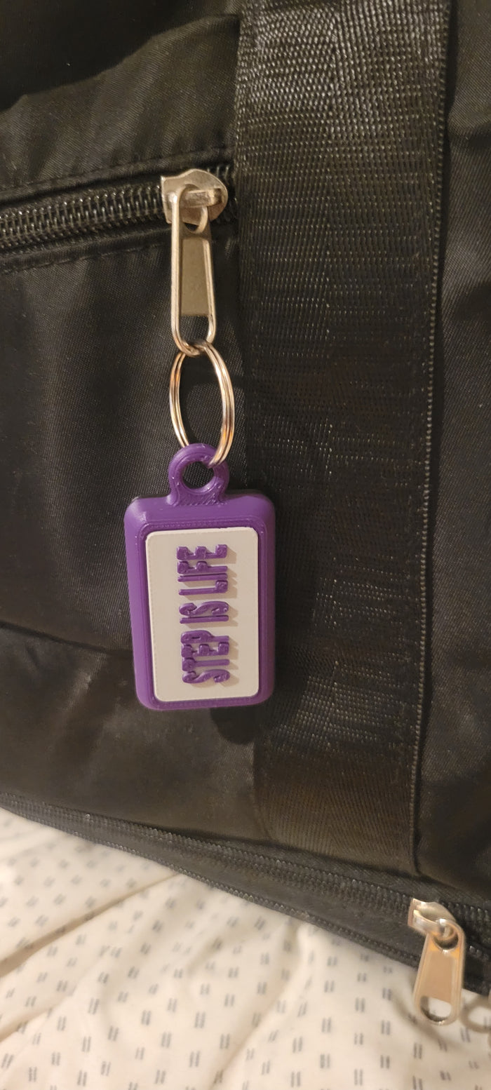 A purple and white rectangular keychain or bag tag with the words 'STEP IS LIFE' in purple, attached to a black duffel bag with a gold zipper pull.