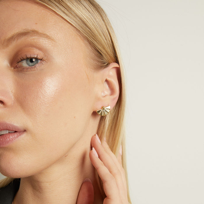 Model. A close-up lifestyle shot of a woman wearing gold fan stud earrings on white background.
