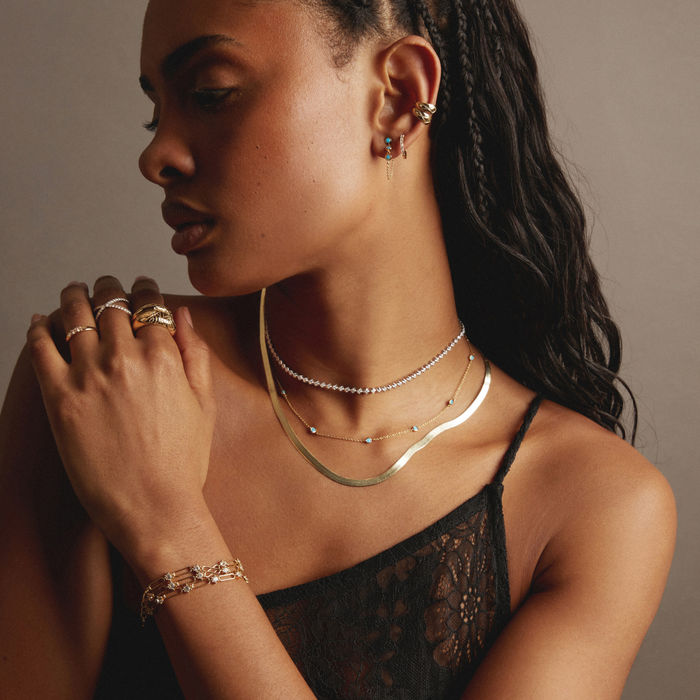 Model. Woman wearing multiple necklaces, earrings, and rings on a neutral background