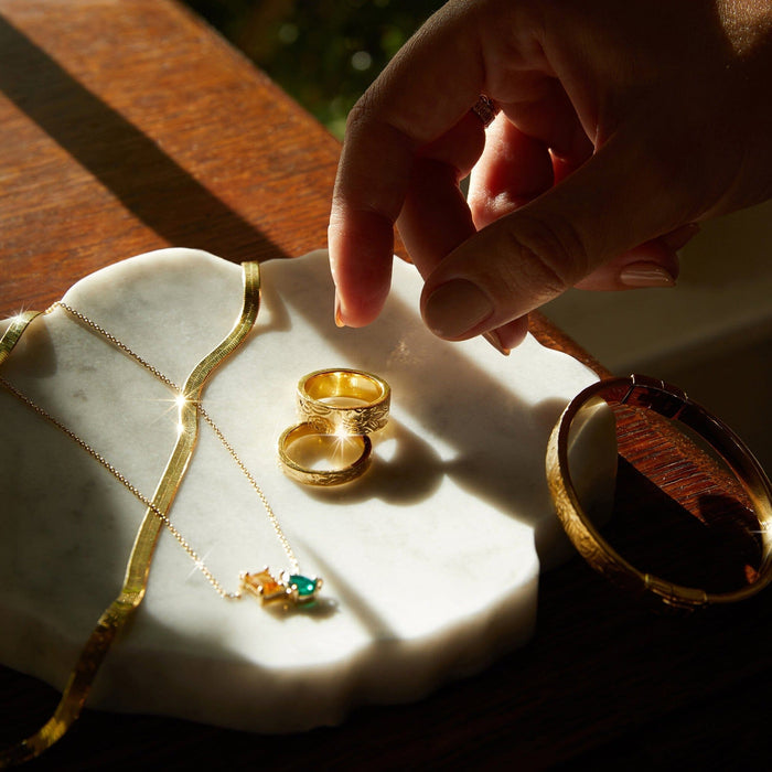 Flatlay Model Close-up of a woman's hand reaching toward gold rings and a necklace resting on a white marble surface."