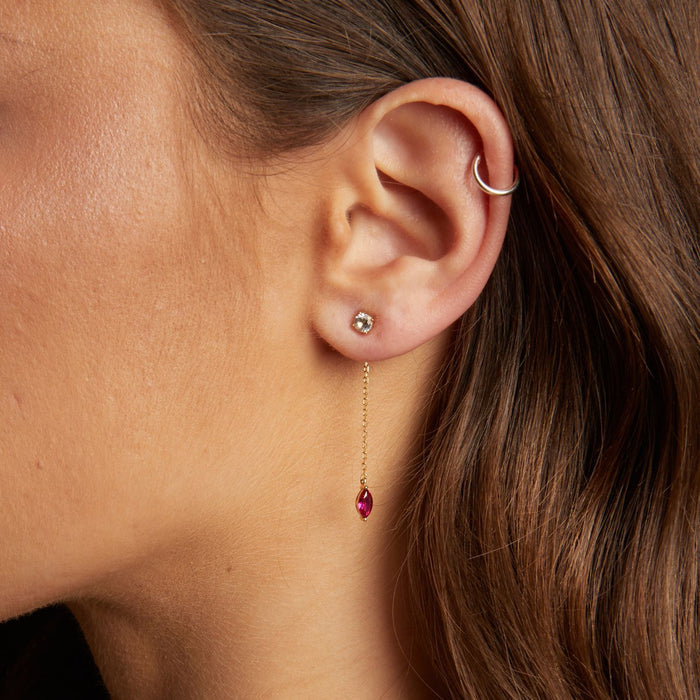 Model. Close-up of an ear wearing a gold earring with a red gemstone and a silver hoop.