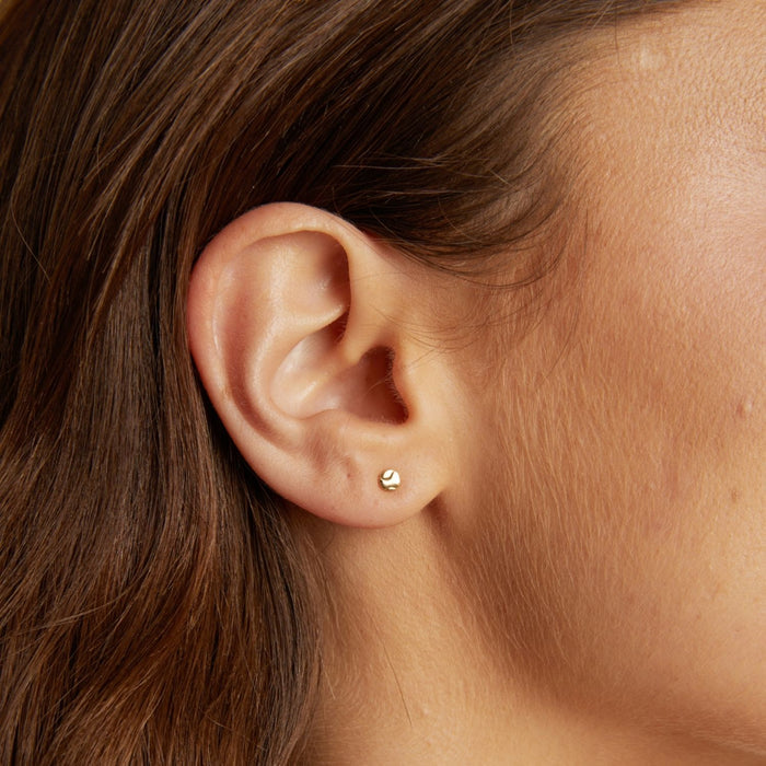 Model. Close-up of an ear wearing a gold stud earring with hair partially covering the side.