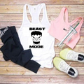 A flat-lay of athletic apparel and accessories on a wooden surface: a white racerback tank top with a black graphic and the words 'BEAST MODE,' a pink sports bra, black leggings, a pink Nike-branded tumbler, and white sneakers.