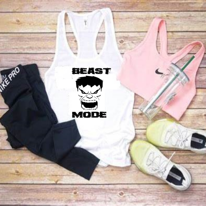 A flat-lay of athletic apparel and accessories on a wooden surface: a white racerback tank top with a black graphic and the words 'BEAST MODE,' a pink sports bra, black leggings, a pink Nike-branded tumbler, and white sneakers.