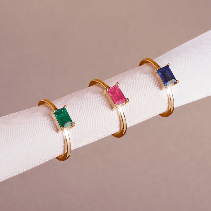 Model Close-up of three gold rings with green, pink, and blue baguette gemstones resting on a white display