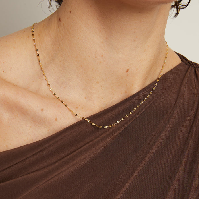 Model. Gold necklace on a person wearing a brown garment against a neutral background