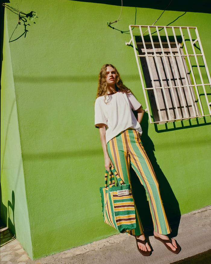 Harly Tee in white cotton, Pasha Trouser in green/yellow/brown striped cotton-linen, Chase Brown Flip Flop, and Sol Green/Yellow Crochet Beach Tote.