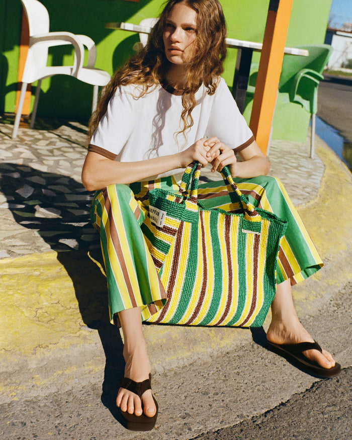 Harly Tee in white cotton, Pasha Trouser in green/yellow/brown striped cotton-linen, Chase Brown Flip Flop, and Sol Green/Yellow Crochet Beach Tote.