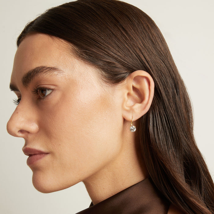 Model. Close up of a woman wearing gold hoop earring with white gemstone on neutral background.