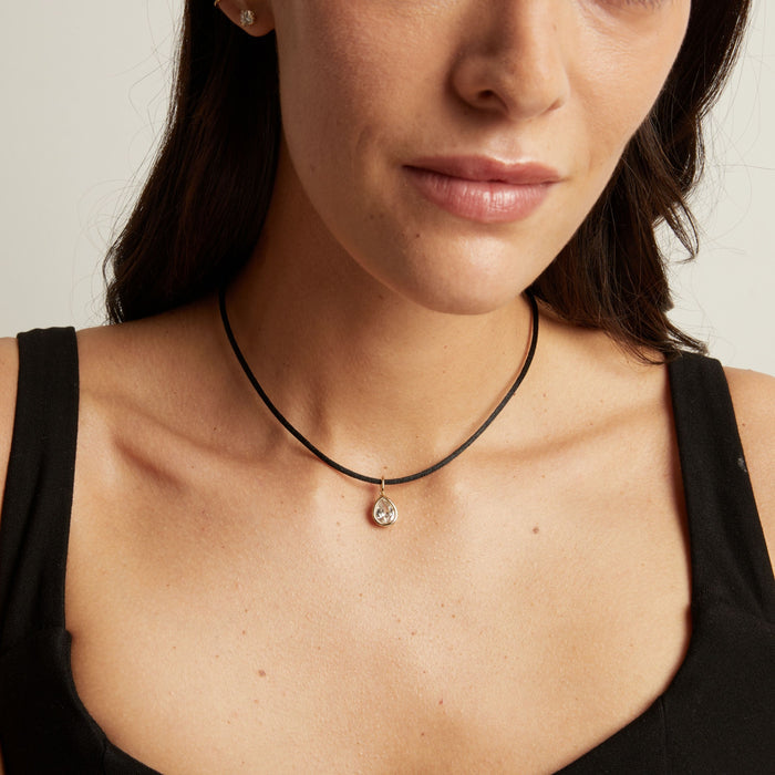 Model. A close-up of a woman wearing a delicate black cord necklace with a small silver-toned pendant featuring a clear teardrop-shaped stone.
