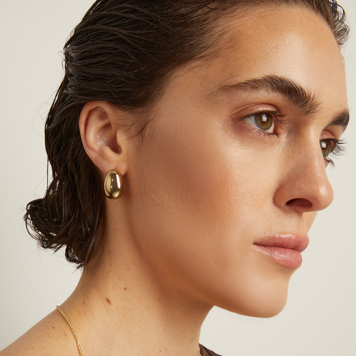 Model. Close-up of a person wearing gold earrings and a necklace against a neutral background