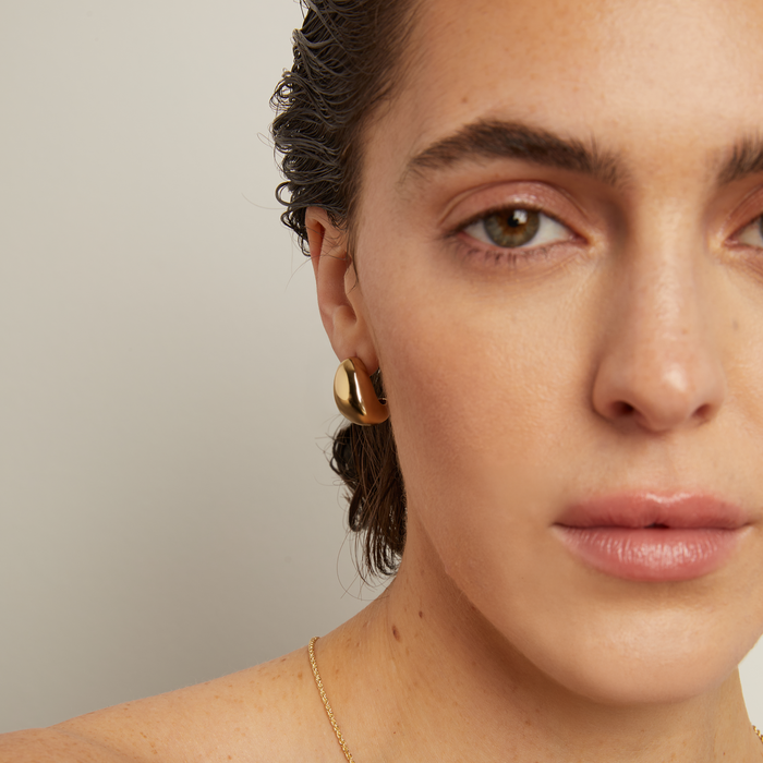 Model. Close-up of a woman wearing gold earrings and a necklace against a neutral background