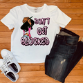 A white graphic t-shirt featuring a cartoon woman and the text 'DON'T GET BLOCKED' in pink and black, styled in a flat lay with dark wash jeans and white sneakers on a wooden floor.