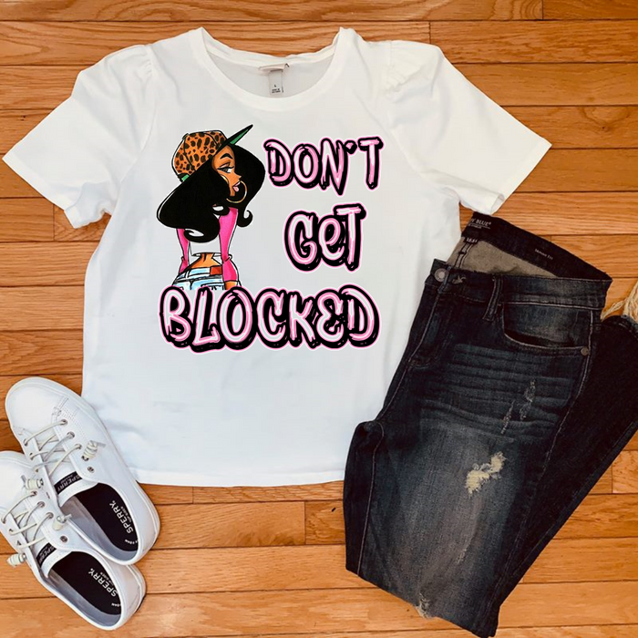 A white graphic t-shirt featuring a cartoon woman and the text 'DON'T GET BLOCKED' in pink and black, styled in a flat lay with dark wash jeans and white sneakers on a wooden floor.