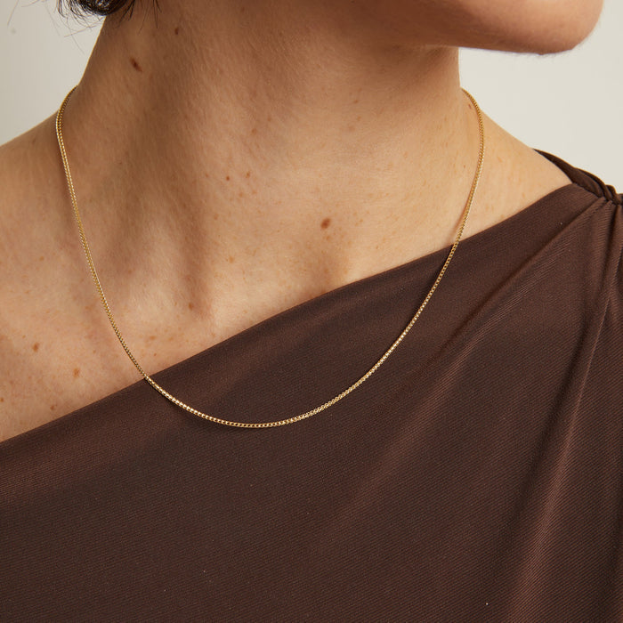 Model. Gold necklace on a person wearing a brown top against a neutral background