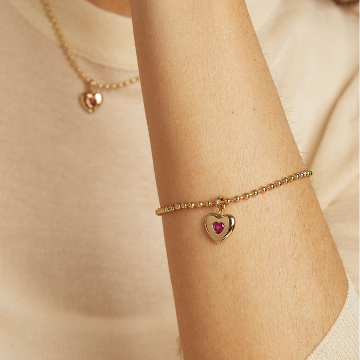 Model. Gold bracelet with a heart-shaped charm on a person's wrist against a neutral background