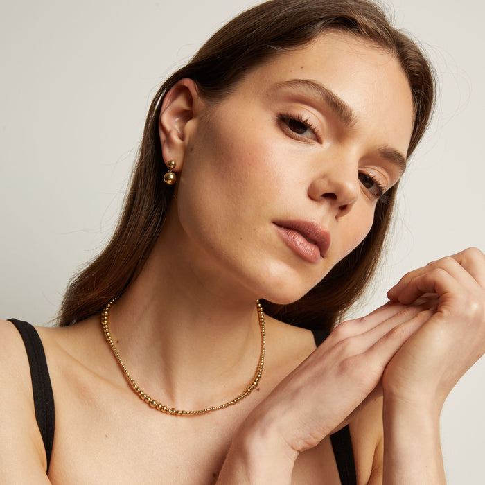 Model. Woman wearing gold jewelry including earrings and a necklace against a neutral background.