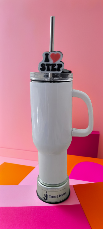 A white insulated tumbler with a handle and clear lid, featuring a metal straw and a decorative 'I <heart> STEP' topper. The tumbler is set against a pink background with colorful geometric shapes on a surface.