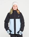 A young person wearing a white beanie, matching gloves, and the Volcom Kids’ Sass’N’Frass Insulated Jacket—crafted from recycled polyester with PFAS-free DWR in black and light blue—stands against a plain white background, looking at the camera with a slight smile.