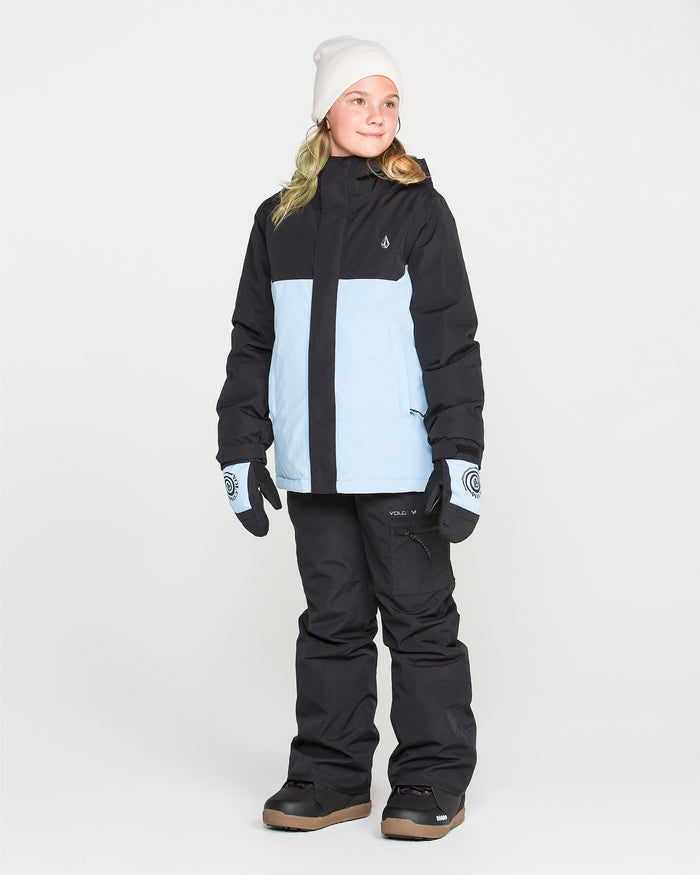 A child wearing the Volcom Kids’ Sass’N’Frass Insulated Jacket in black and light blue, paired with black snow pants, gloves, brown boots, and a white beanie stands against a plain white background, looking slightly to the side and smiling.