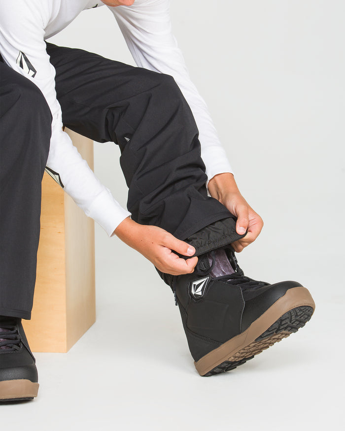 A person wearing Volcom Kids’ Youth Freakin Chino Insulated Pants in black, featuring an adjustable waist, pairs them with a white long-sleeve shirt and sits on a wooden bench, tucking the pant legs into black winter boots with brown soles.