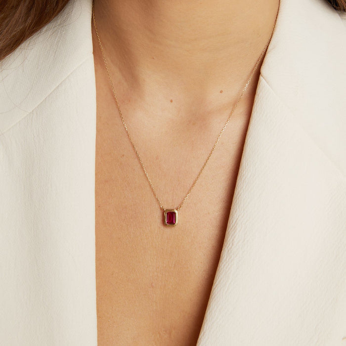 Model. Gold necklace with a red gemstone worn by a person in a white outfit.