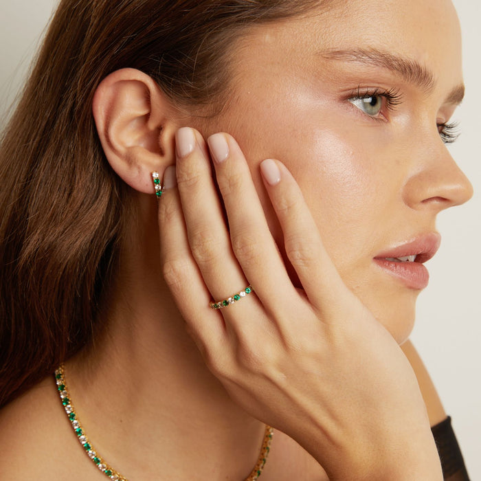 Close-up of a woman wearing emerald green jewelry including earrings and a ring, with a neutral background.