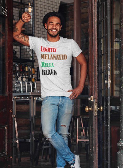 A smiling man wearing a white graphic t-shirt with the text 'LIGHTLY MELANATED MELLA BLACK' in red, brown, green, and black fonts, paired with distressed blue jeans, standing in a doorway of what appears to be a bar or restaurant.