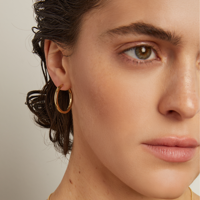 Model. Close-up of a woman's face wearing a gold hoop earring against a neutral background