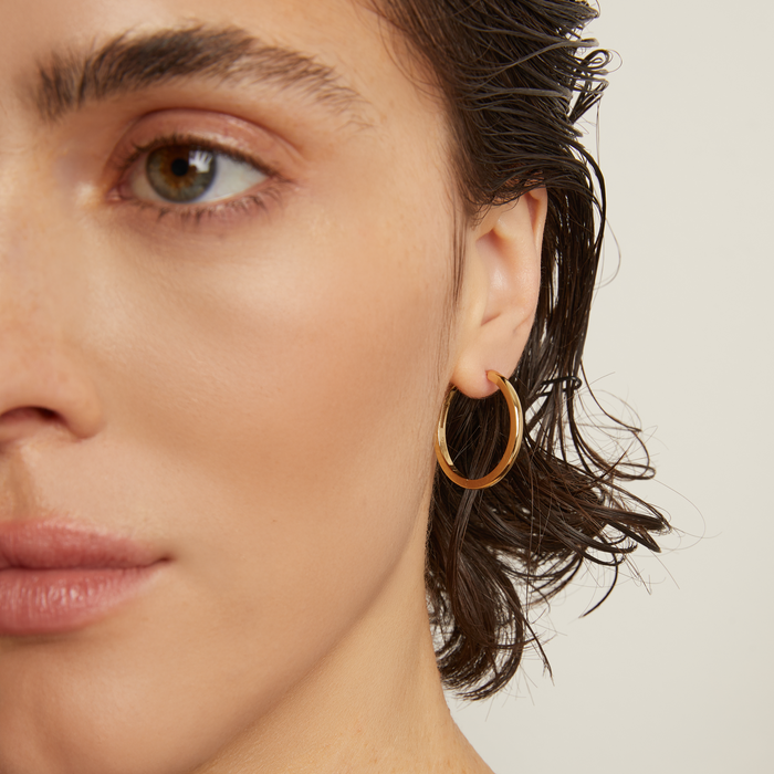 Model. Close-up of a person wearing gold hoop earrings with a neutral background