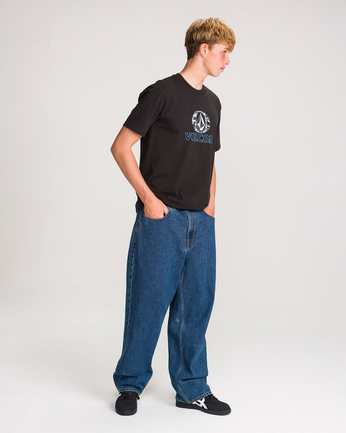 A person stands sideways against a plain white background, hands in their pockets, wearing loose-fit blue jeans—the Men’s Billow Denim Pants by Volcom—paired with a black graphic T-shirt and black sneakers. Their short hair is light brown.