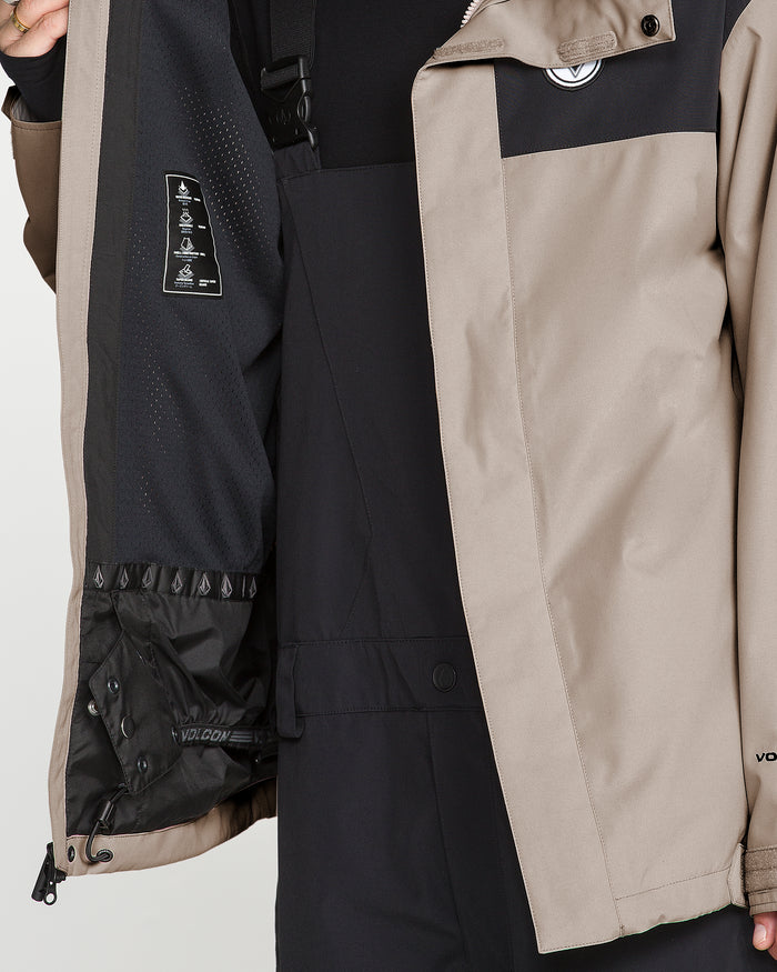 A person models the Volcom Men’s Buckthreeeighty Jacket in tan, holding it open to show the black lining with labels and a black inner section featuring V-Science 2-Layer construction. The image focuses on the torso and part of the arm, emphasizing the jacket’s PFAS-free DWR finish.