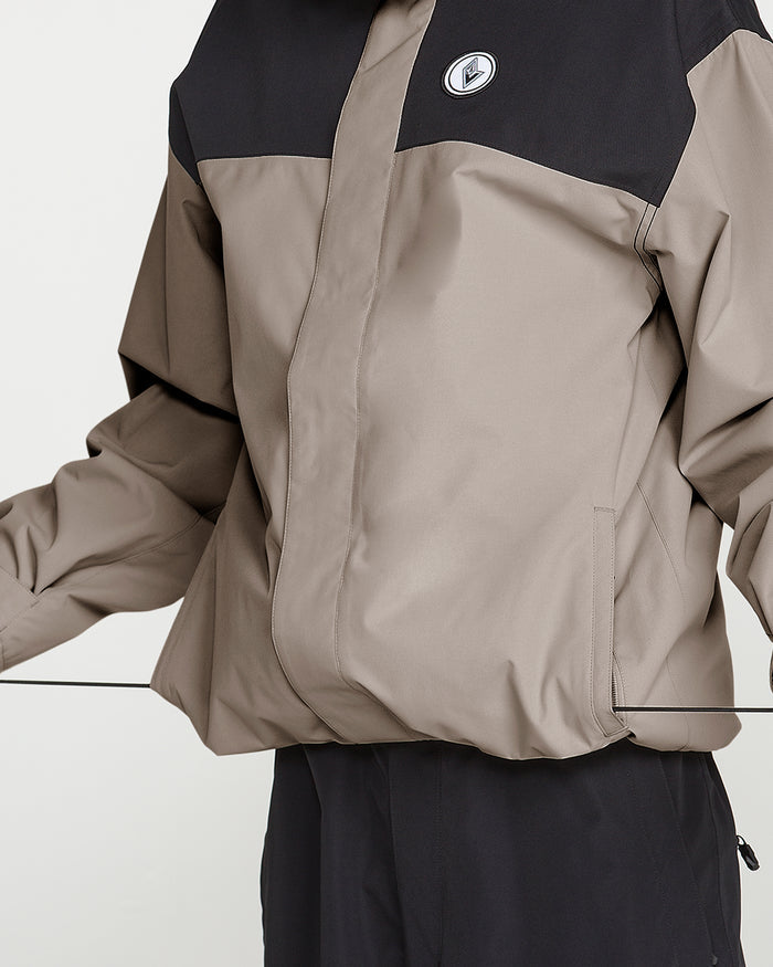 A man wearing the Volcom Men’s Buckthreeeighty Jacket in beige and black, featuring a circular logo on the chest, PFAS-free DWR coating, and V-Science 2-Layer technology. The image shows only his torso and part of his arms against a plain white background.