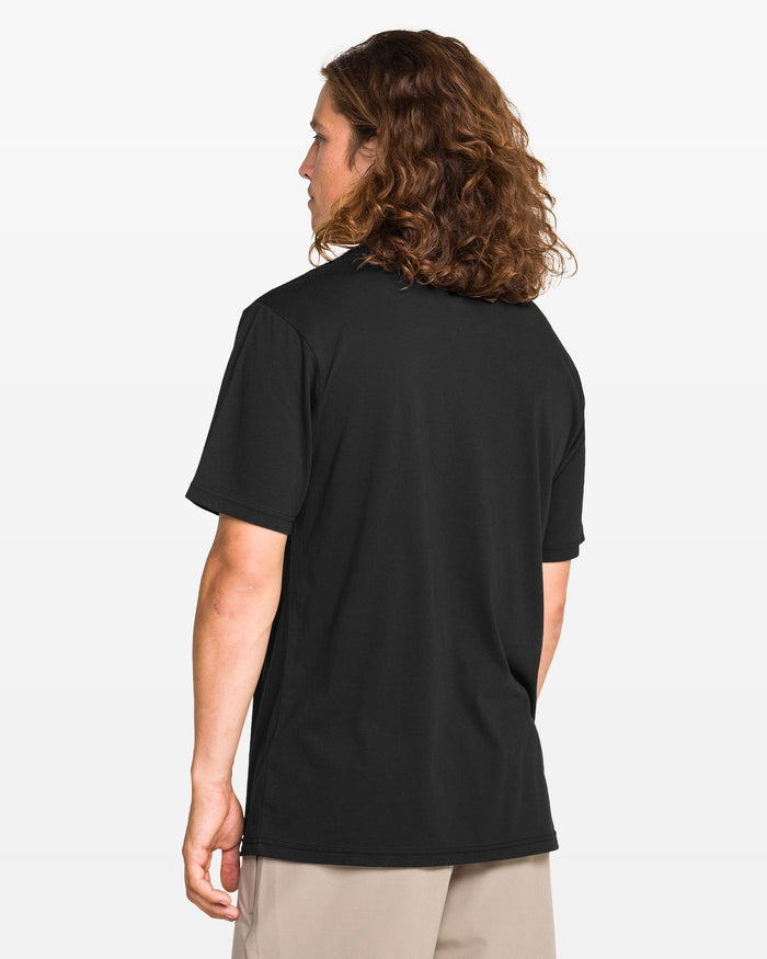A person with long brown hair, wearing a black Volcom Men's Enterprise Short Sleeve Tee in a modern fit and beige pants, stands with their back to the camera against a plain light background.