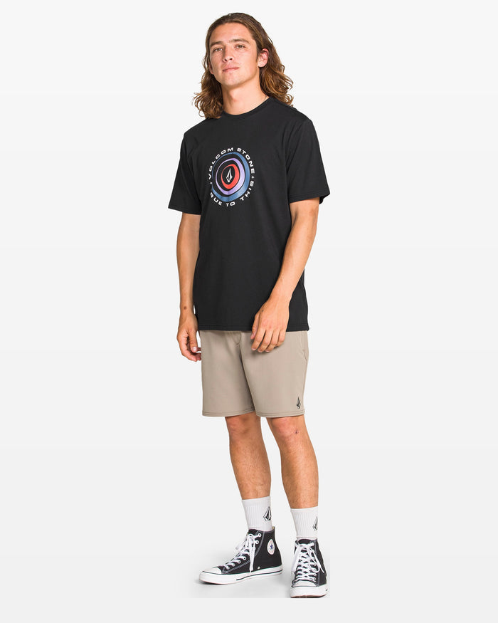 A young man with long hair faces forward, wearing the Volcom Men's Enterprise Short Sleeve Tee in black, paired with beige shorts, white socks, and black high-top sneakers against a plain white background.