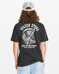 A person with long hair, wearing a white mesh cap and the Volcom Men's Eternal Short Sleeve Tee in black with a soft-hand reaper graphic, plus light blue shorts, is seen from behind against a plain backdrop.