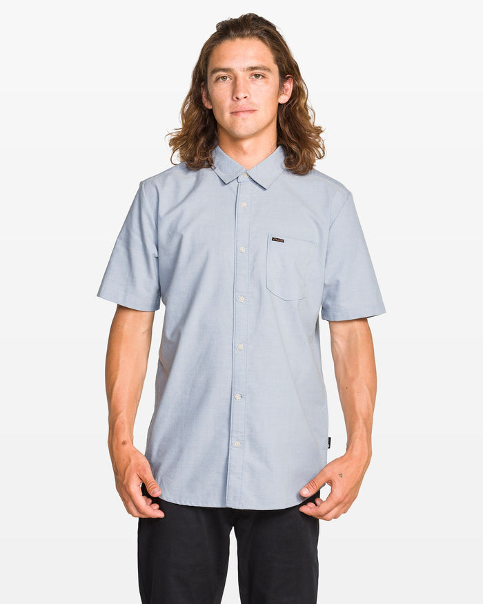 A young man with long brown hair wears a Volcom Men's Everett Oxford II Shirt in light blue and black pants, standing facing forward against a white background to showcase its classic fit.