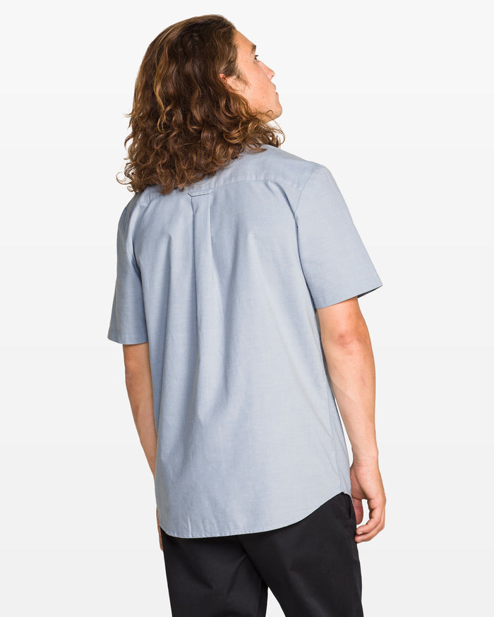 A person with long, wavy brown hair faces away, wearing a light blue Volcom Men's Everett Oxford II Shirt in a classic fit, paired with black pants against a plain white background.