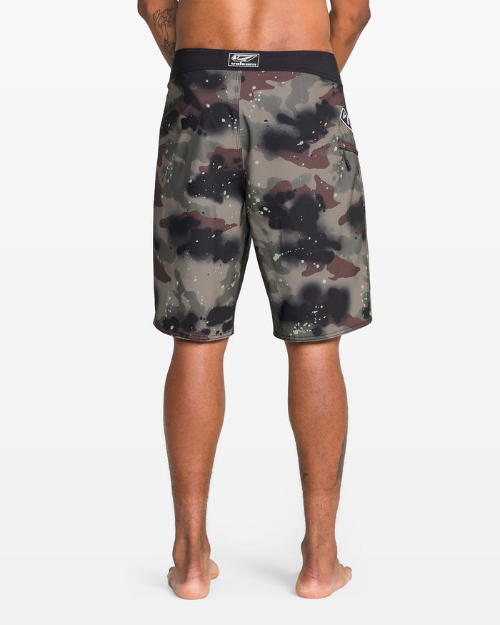 A person stands barefoot facing away, wearing Volcom Men's Fomenter Mod board shorts with knee-length camouflage print, 4-way stretch Stone Shield fabric, and a black waistband. Only their legs and lower back are visible against a white background.
