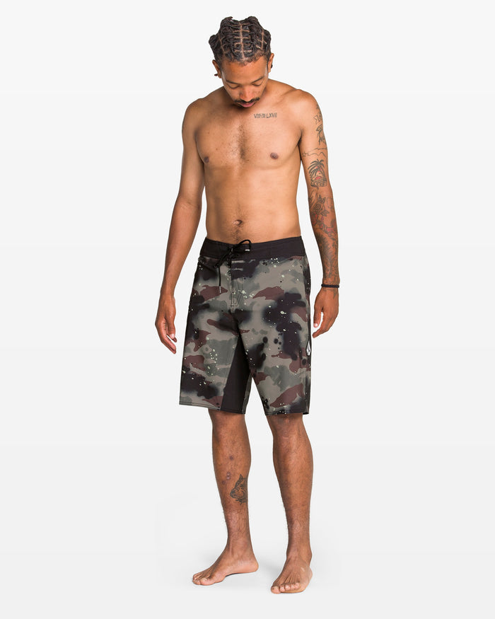 A shirtless, tattooed man with braided hair stands barefoot in Volcom's Men's Fomenter Mod knee-length camo board shorts made from REPREVE recycled fibers with a drawstring waist, looking down against a plain white background.