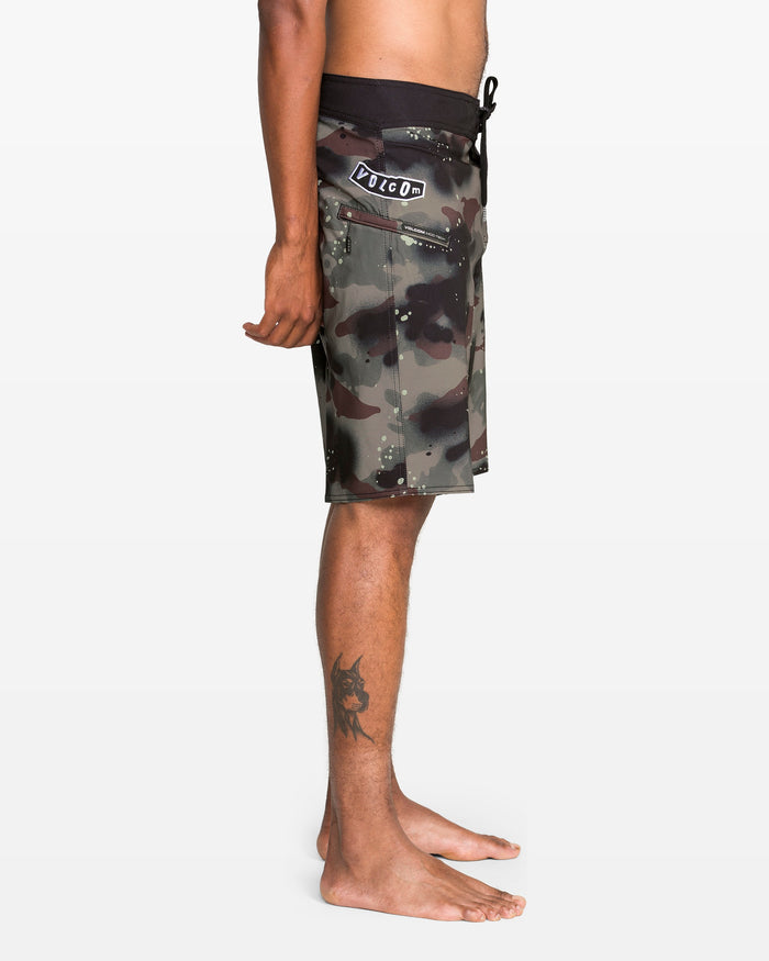 A person stands barefoot in profile, wearing Volcom Men's Fomenter Mod board shorts with a camouflage print, 4-way stretch Stone Shield fabric, black waistband with Pico patch, and a visible tattoo on their lower right leg.