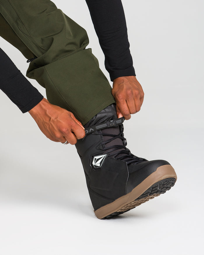 A person wearing Volcom Men’s Freakin Snow Chino Pants in green and a black shirt tightens the laces on a black snowboarding boot with a brown sole against a plain light background.