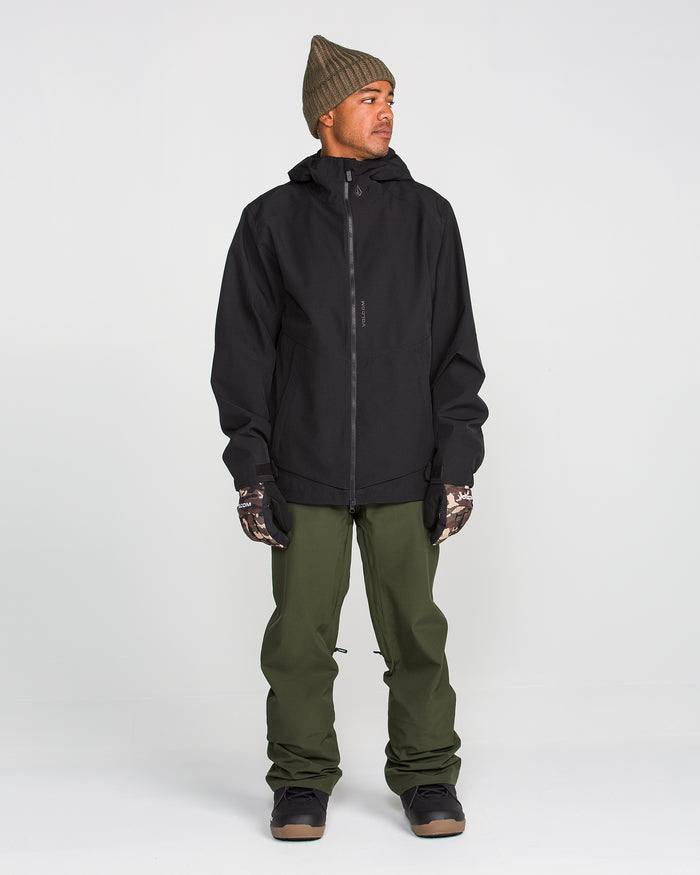A person stands against a plain background, wearing a black hooded jacket, Volcom Men’s Freakin Snow Chino Pants in olive green, camouflage gloves, dark boots, and a brown knit beanie. The waterproof and breathable Volcom pants complete the look as the person gazes to the side with hands by their sides.