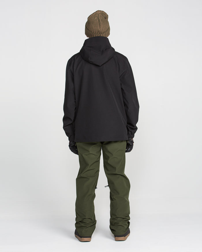 A person stands facing away, wearing a brown beanie, black hooded jacket, Volcom Men’s Freakin Snow Chino Pants in waterproof, breathable green, and gloves against a plain white background.
