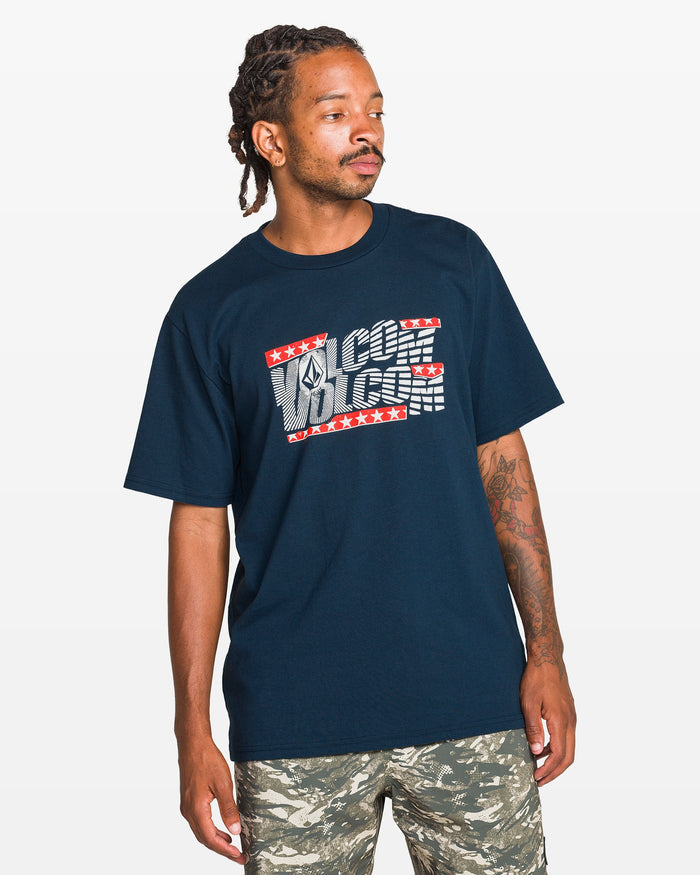 A man with dreadlocks wears a navy blue Volcom Men's Glory Daze Short Sleeve Tee and camouflage shorts, standing against a plain white background and looking slightly to his left.