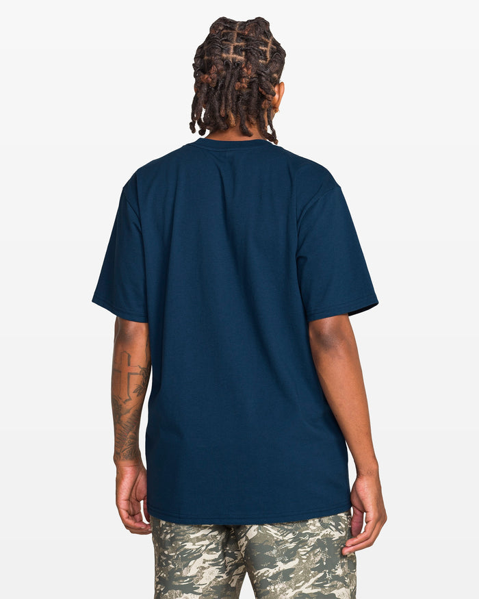 A person with dreadlocks is seen from the back wearing Volcom’s Men’s Glory Daze Short Sleeve Tee from the Froth of July Collection paired with camo shorts. A tattoo is visible on their left arm, against a plain white background.
