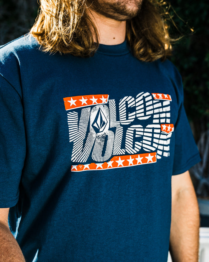 A person with long hair wears the Volcom Men's Glory Daze Short Sleeve Tee, a blue shirt from the Froth of July Collection, featuring a bold white logo with orange stars and stripes. The image is brightly lit and highlights the shirt’s design.