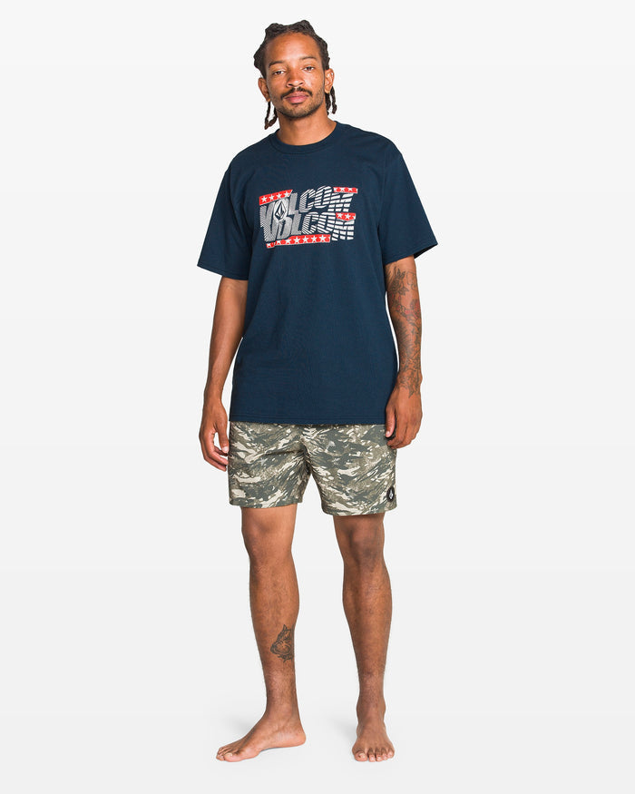 A man stands barefoot against a white background, wearing a navy blue Volcom Men's Glory Daze Short Sleeve Tee from the Froth of July Collection with a graphic logo and green camo shorts.