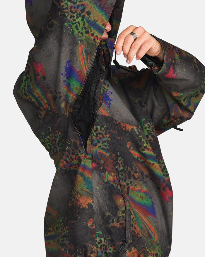 A person wearing the Volcom Men’s Hossegor 20K Pullover, a colorful, patterned jacket with animal print details, is unzipping a side zipper near the armpit area. The hood is up, partially obscuring their face. This pullover features a recycled V-Science shell for sustainable style and performance.